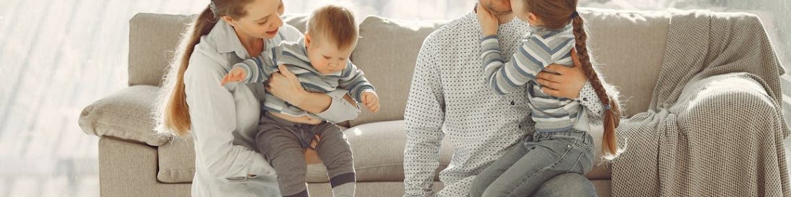 Happy parents with children on knees in casual clothes sitting on floor and playing with kids while spending time together in living room near panoramic window